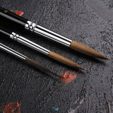 Fuumuui & Svetlin Sofroniev Master's Set Ultimate Collection professionelle Künstlerfarbenpinsel mit drei runden Pinseln mit natürlichen braunen Borsten, silbernen Zwingen und schwarzen Griffen auf einer dunklen, strukturierten Oberfläche.