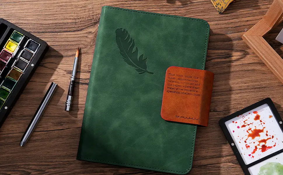 Green leather Fuumuui watercolor sketchbook with 100% cotton cold press paper. Professional set includes a travel watercolor brush, watercolor paint set with pans, and travel palette on a wooden artist desk.