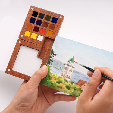 Set di acquerelli in legno premium Fuumuui con 15 colori selezionati a mano e una tavolozza di miscelazione integrata, mostrato aperto mentre un artista dipinge un paesaggio dettagliato.
