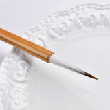 Fuumuui Chinese Calligraphy Wolf Sheep Mix Brush with natural bamboo handle and pointed white and brown hair tip, resting on a white decorative plate.