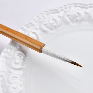 Fuumuui Chinese Calligraphy Wolf Sheep Mix Brush with natural bamboo handle and pointed white and brown hair tip, resting on a white decorative plate.