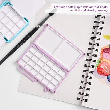 Mini palette de mélange d'aquarelle Fuumuui en violet doux, ouverte pour révéler 24 godets et plateaux de mélange doubles pour la peinture portable, montrée avec un pinceau Fuumuui et une palette bleue sur un carnet de croquis d'art.