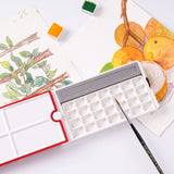 Palette d'aquarelle de voyage 28 godets Fuumuui ouverte, présentant son couvercle rouge rotatif à 360°, ses multiples plateaux de mélange et sa conception compacte avec un emplacement pour pinceau. Un pinceau d'aquarelle repose à proximité, entouré de fournitures d'art Fuumuui et d'œuvres d'art aquarellées terminées.