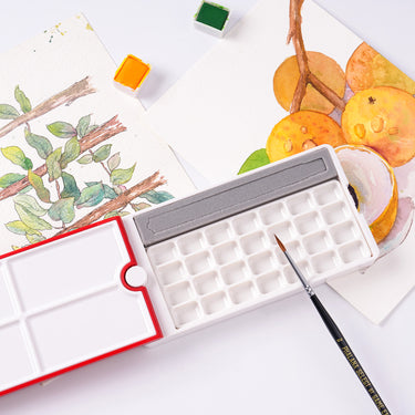 Palette d'aquarelle de voyage 28 godets Fuumuui ouverte, présentant son couvercle rouge rotatif à 360°, ses multiples plateaux de mélange et sa conception compacte avec un emplacement pour pinceau. Un pinceau d'aquarelle repose à proximité, entouré de fournitures d'art Fuumuui et d'œuvres d'art aquarellées terminées.