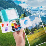 Boîte de palette d'aquarelle pliante double couche Fuumuui, un plateau de peinture bleu compact avec 24 godets et 7 zones de mélange, tenu par un artiste peignant un paysage extérieur pittoresque, soulignant sa conception portable pour les voyages et l'utilisation en studio avec Fuumuui Art Supplies.