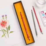 Fuumuui Art Supplies: pennelli per acquerello artigianali in pelo di capra rosso con manici rossi e verdi, uno presentato in una scatola decorativa, accanto a un vivace dipinto di fiori e una tavolozza di acquerelli.