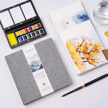 Fuumuui Watercolor Book (7.6×7.6" Cold-Press 100% Cotton) displayed with Fuumuui art supplies, including a watercolor palette and brush, featuring a vibrant autumn tree painting.
