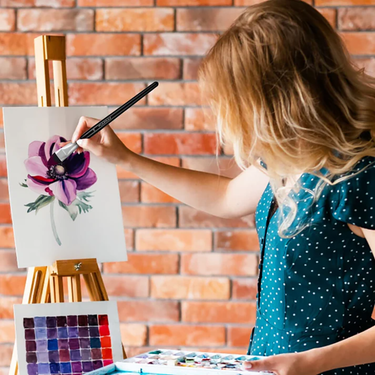Fuumuui 8pcs Professional Squirrel Watercolor Brushes are demonstrated as a woman paints a purple flower on an easel.