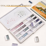 Set di acquerelli semi-solidi opachi da 24 colori Fuumuui con colori tenui, esposto aperto nella sua custodia di metallo bianca con godet rimovibili, accanto a un album da schizzi per acquerelli. Questo articolo per belle arti Fuumuui ¨¨ ideale per principianti e artisti.