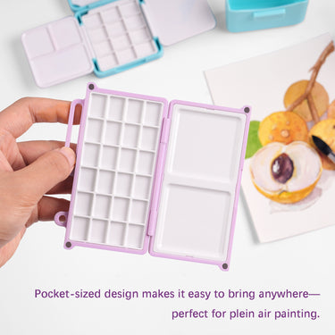 Main tenant une mini palette de mélange d'aquarelle lavande Fuumuui ouverte avec 24 godets et plateaux doubles, parfaite pour les fournitures d'art de plein air portables.