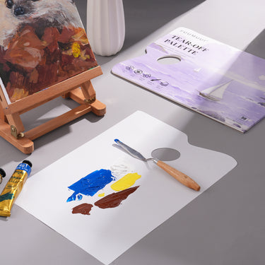 Fuumuui Einweg-Farbpaletten-Papierblock, 22,9 x 30,5 cm, mit 30 Blatt abreißbarem Papier, das Acryl- und Ölfarben hält, die mit einem Spachtel gemischt wurden, neben einer Staffelei des Künstlers. Essentielles Fuumuui-Künstlermaterial.