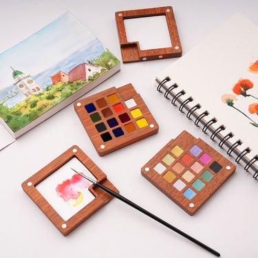 Set di acquerelli perlati Fuumuui, con 15 colori iridescenti in un'elegante tavolozza di legno magnetica, esposto con un vassoio di miscelazione, un pennello e opere d'arte ad acquerello su un album da disegno, parte di Fuumuui Art Supplies.