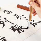 Close-up of a hand holding a Fuumuui Chinese Calligraphy Wolf Sheep Mix Brush, actively writing elegant Chinese characters on textured paper with black ink.