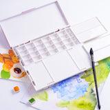 Boîte de palette de peinture hermétique Fuumuui ouverte, présentant ses 28 godets vides et ses plateaux de mélange de couvercle pratiques. Un pinceau d'aquarelle, de petits cubes de peinture et des peintures à l'aquarelle finies accompagnent la palette, idéales pour les fournitures d'art d'aquarelle, de gouache et d'acrylique.