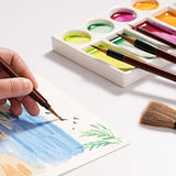 Hand uses a Fuumuui professional synthetic goat mixed hair watercolor detail brush to paint a landscape, with a vibrant watercolor palette and other Fuumuui water color paint brushes from the set visible.