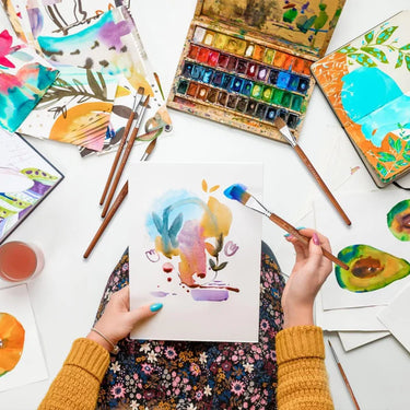 Artist using Fuumuui professional watercolor brushes, including a flat brush, to paint abstract art with a colorful palette and other Fuumuui round and flat brushes visible.
