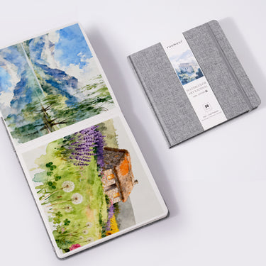 Fuumuui Watercolor Book 7.6x7.6 Cold-Press 100% Cotton, with one art journal open showcasing vibrant watercolor paintings of mountains and a rustic cottage, and another Fuumuui Art Supplies journal closed with a grey fabric cover and elastic band.
