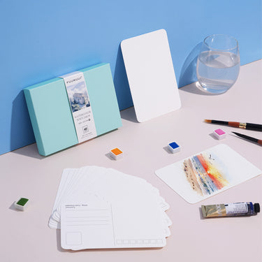 FUUMUUI Aquarellpostkarten mit Umschlag, 100% Baumwolle & kaltgepresst, mit einer bemalten Postkarte neben leeren Karten, Aquarellfarben, Pinseln und anderem FUUMUUI-Kunstbedarf, bereit für die Gestaltung.