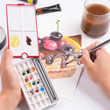 Palette d'aquarelle de voyage 28 godets Fuumuui ouverte avec couvercle rouge rotatif à 360° et plateaux de mélange, montrant sa conception compacte et son emplacement pour pinceau, tandis qu'une personne utilise des fournitures d'art Fuumuui pour peindre un gâteau à l'aquarelle.