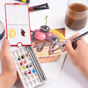 Palette d'aquarelle de voyage 28 godets Fuumuui ouverte avec couvercle rouge rotatif à 360° et plateaux de mélange, montrant sa conception compacte et son emplacement pour pinceau, tandis qu'une personne utilise des fournitures d'art Fuumuui pour peindre un gâteau à l'aquarelle.