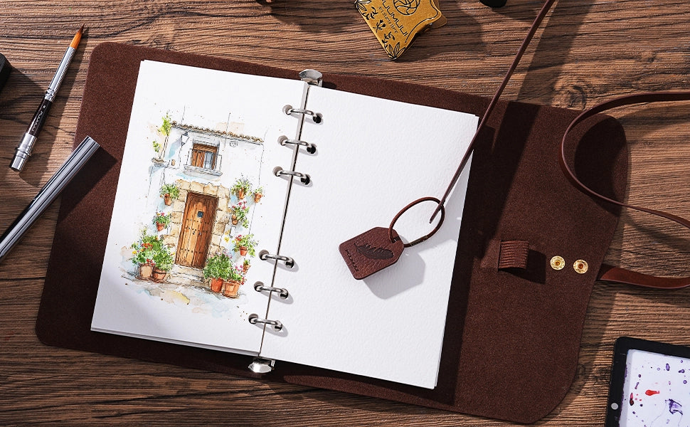 Open leather watercolor sketchbook with 100% cotton cold press paper and a silver travel pocket brush. A ceramic palette with watercolor paint sits beside a professional artist's sketch on a rustic wooden desk.