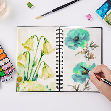 Fuumuui Watercolor Sketchbook – Spiral Bound & 100% Cotton, open on a white table, showcasing vibrant watercolor paintings of yellow and blue flowers, with a hand actively painting using Fuumuui art supplies and brushes nearby.