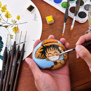 A hand using a Fuumuui fine detail brush to paint a yellow circle on a small rock, surrounded by other painted brushes.