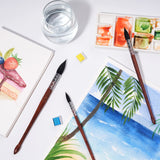 Pennelli a punta tonda in pelo di scoiattolo naturale FUUMUUI per pittura ad acquerello, esposti accanto a una tavolozza, acqua e opere d'arte finite vivaci che raffigurano un dessert colorato e una serena scena di spiaggia. Forniture artistiche Fuumuui essenziali.