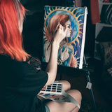 Artista che utilizza un pennello professionale in martora per acquerello Fuumuui dal set di 7 pezzi per dipingere un ritratto colorato su un cavalletto, con una tavolozza di acquerelli accanto.