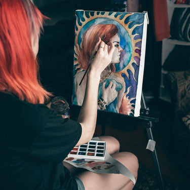 Artista che utilizza un pennello professionale in martora per acquerello Fuumuui dal set di 7 pezzi per dipingere un ritratto colorato su un cavalletto, con una tavolozza di acquerelli accanto.