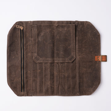 Fuumuui durable dark brown waxed canvas brush roll for art supplies, unrolled to show organized brush slots, a zippered pocket, a flap pocket, and its leather strap with brass buckle closure.