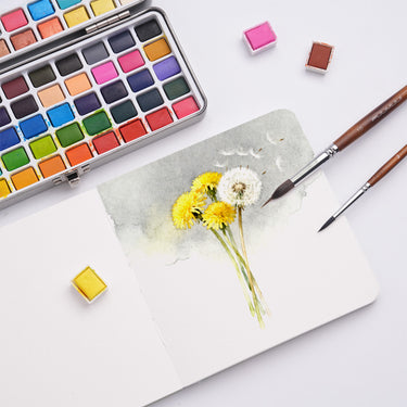 Fuumuui - Blocco per acquerello, 5.5x5.5" Verde Erba, 100% cotone, pressato a freddo, aperto su un vivace dipinto ad acquerello di un dente di leone, accanto a un set di godet per acquerelli e pennelli di Fuumuui art supplies.