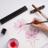 Fuumuui Art Supplies: Eine Hand verwendet einen FUUMUUI Handgefertigten Tierhaarkalligraphiepinsel, um Aquarellblumen zu malen, mit einem anderen FUUMUUI Pinsel mit Magnetkappe, einer Geschenkbox und traditionellen Kunstbedarf sichtbar.