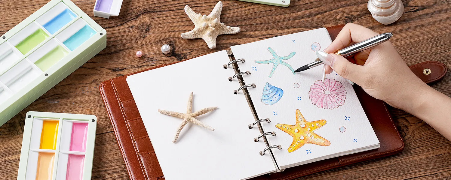 Hand painting a starfish in a journal next to a mint green paint palette filled with pastel colors.