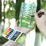 Artista che dipinge un paesaggio all'aperto utilizzando un kit di acquerelli indossabile Fuumuui, con la tavolozza da viaggio a 24 colori sul polso e un pennello.