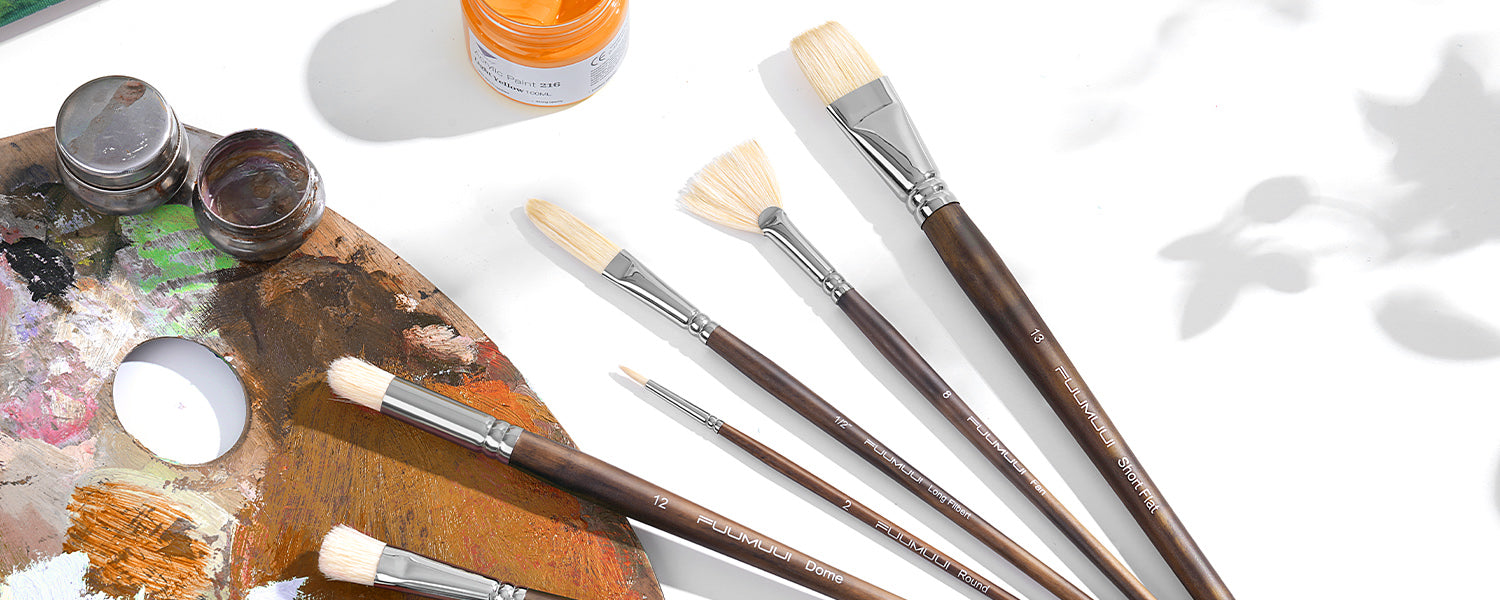 Assorted hog bristle paint brushes laid out on a table with a wooden artist palette and paint jars.