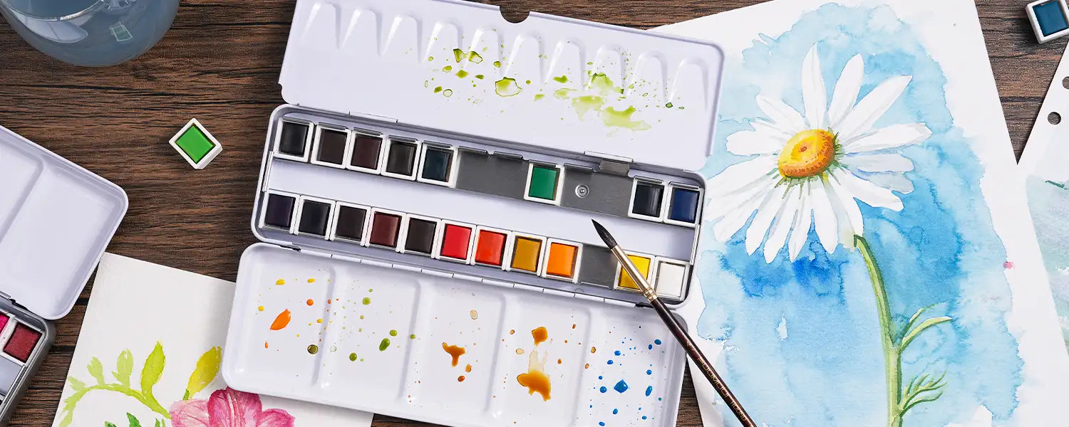 Flat lay of an open watercolor tin set with paint swatches beside a detailed daisy watercolor painting.