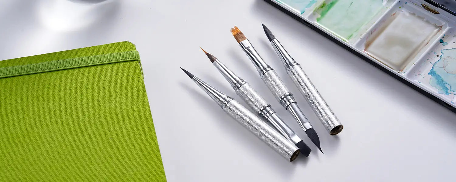 Four disassembled FUUMUUI 3-in-1 Travel Watercolor Brushes lying next to a green sketchbook and a watercolor palette.