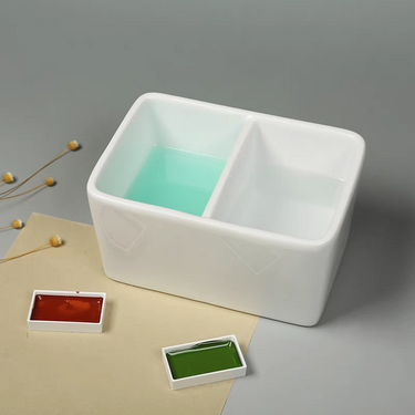 Fuumuui Keramik-Pinselwaschbecken, weiß mit zwei Fächern, die klares und blaues Wasser enthalten, neben roten und grünen Aquarellfarben.